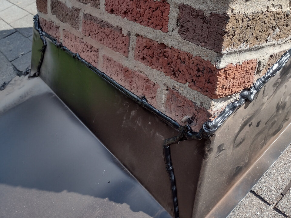 Metal flashing wrapped around the corner of a brick chimney on a shingle roof, with sealant along its top edge.