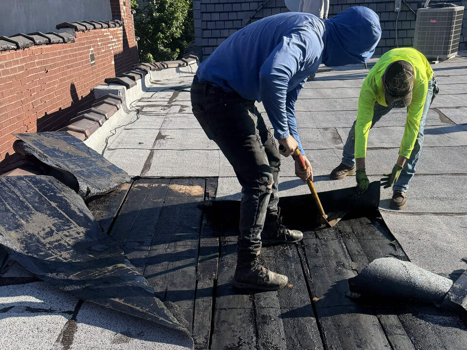 Two roofers removing old roofing layers from a flat roof, exposing the wood plank deck.