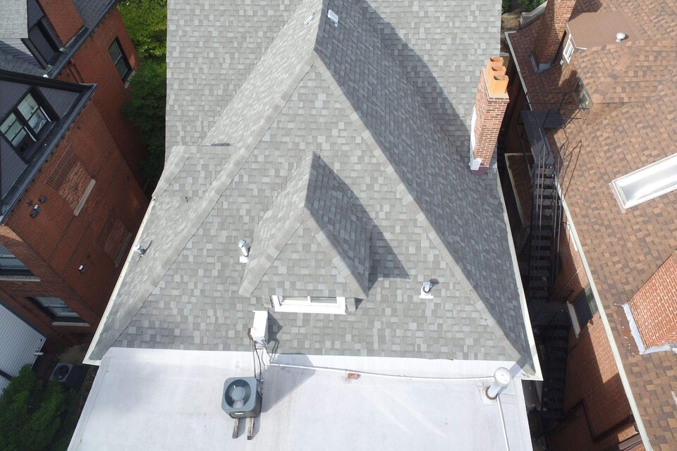 Overhead aerial view of a gray shingle roof with hips, a dormer, roof vents and a brick chimney, joined to a flat lower roof and set between neighboring brick buildings.