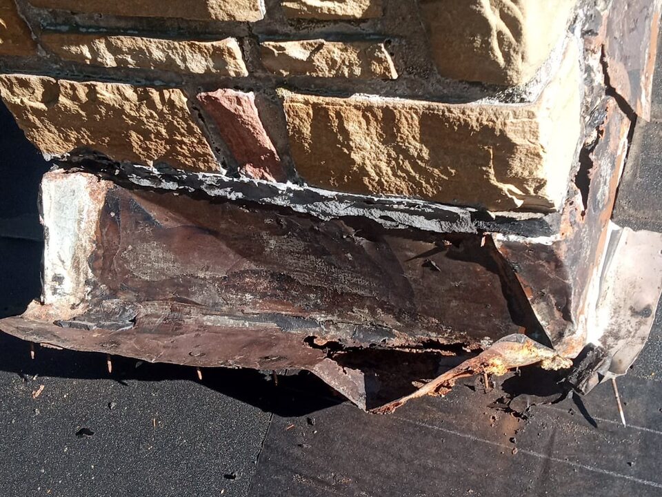 Rusted, torn metal flashing at the base of a stone chimney, with a dark roof surface below.