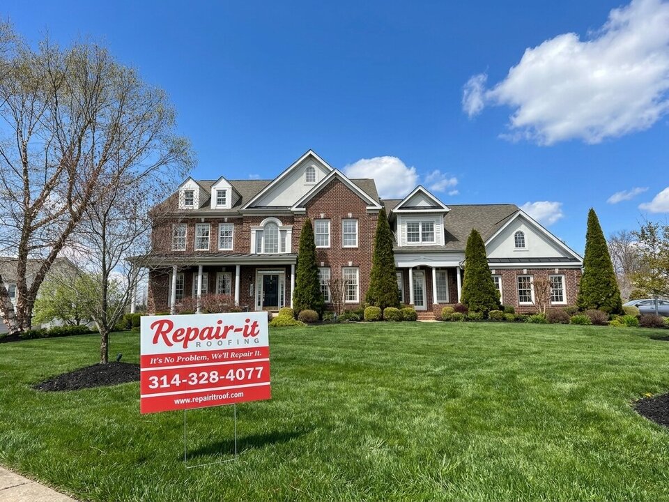 A brick house with a Repair It Roofing yard sign on the front lawn.